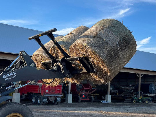 Tractor equipped with a Bale Spear moving multiple round hay bales across a barnyard.